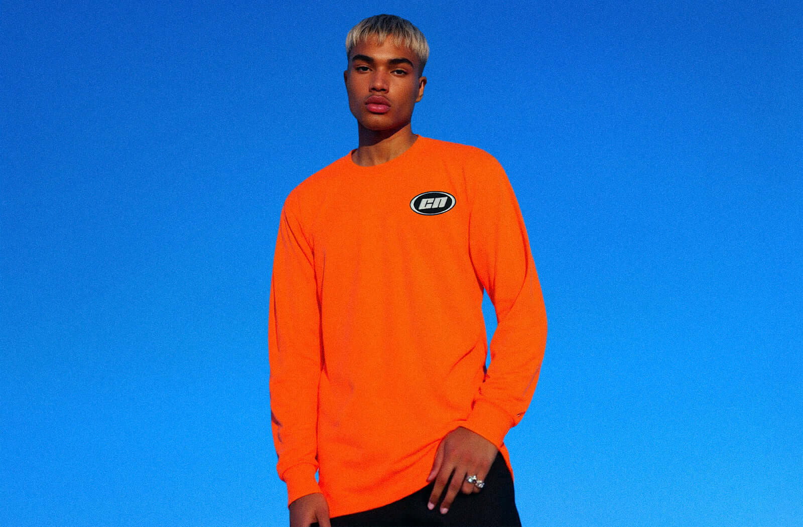 Model wearing a bold orange long-sleeve top against a blue sky, representing Club Neo's stylish collection.