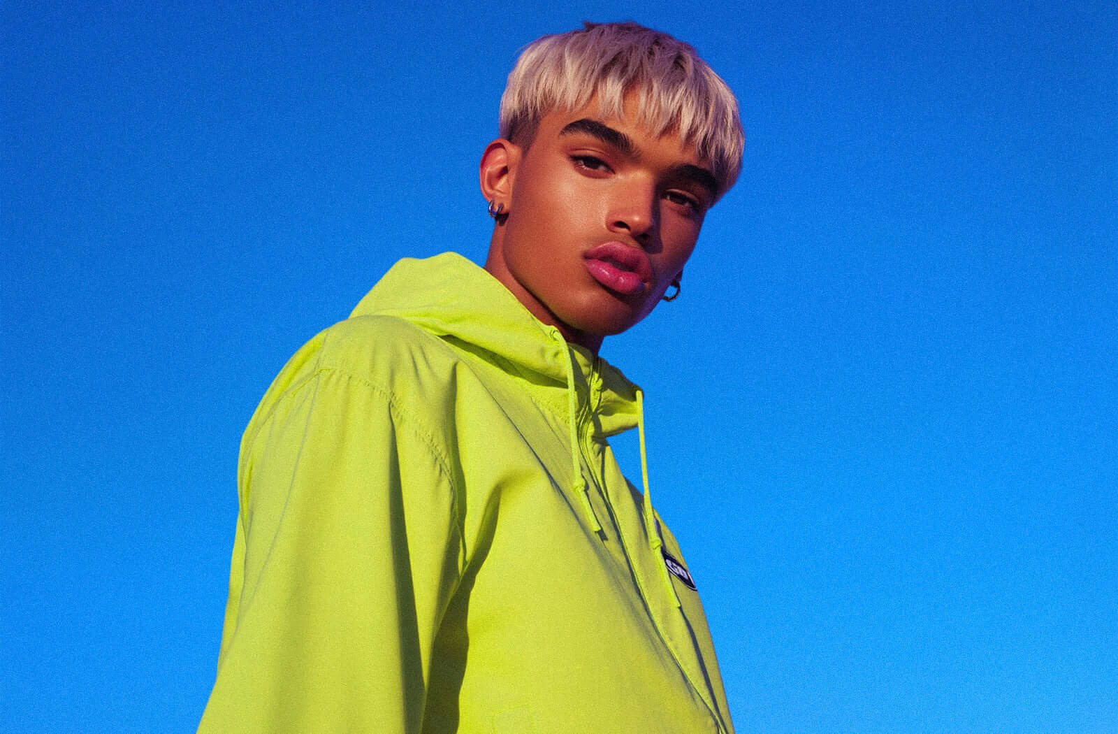 Model wearing a vibrant neon windbreaker against a clear blue sky, showcasing futuristic streetwear style.