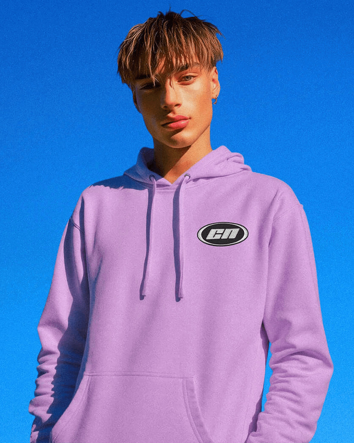 Model wearing a lavender pullover hoodie with embroidered logo against a bright blue sky.