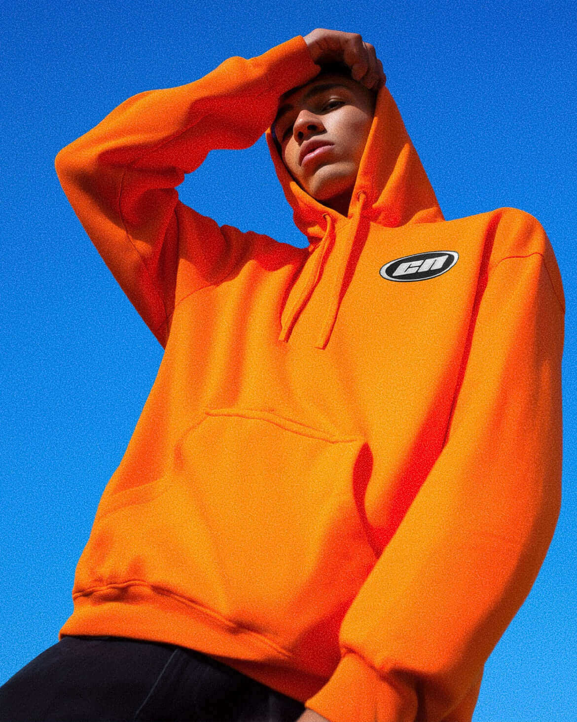 Model wearing neon orange pullover hoodie with adjustable hood and kangaroo pocket against a blue sky.