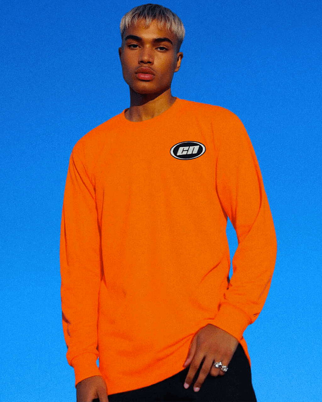 Model wearing a long sleeve neon orange cotton t-shirt with embroidered Club Neo logo against a blue sky.