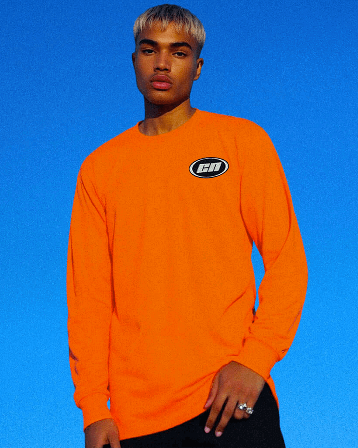 Model wearing a long sleeve neon orange cotton t-shirt with embroidered Club Neo logo against a blue sky.