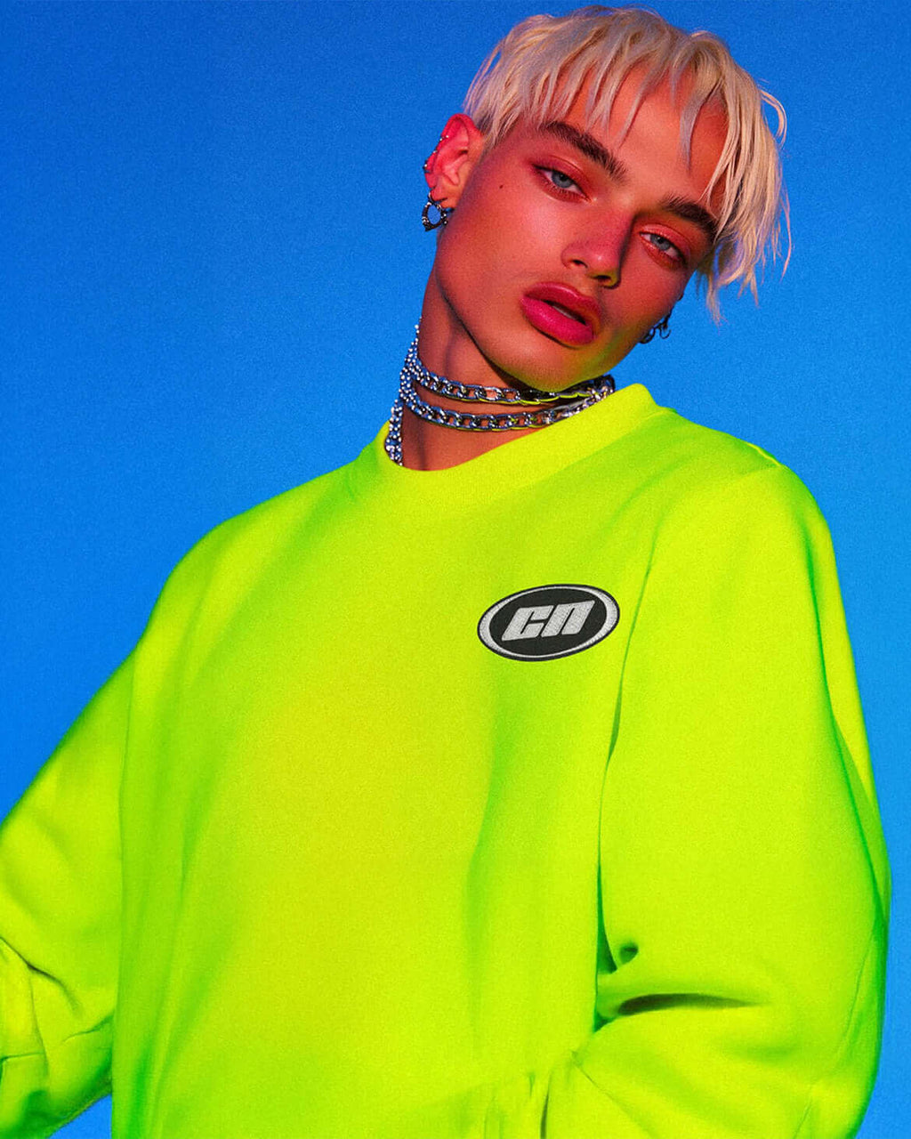 Model wearing a lightweight neon green crewneck sweater featuring an embroidered logo, against a vibrant blue background.