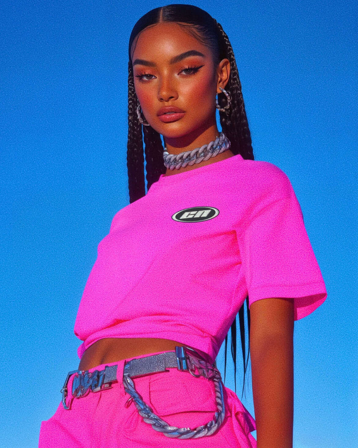 Model wearing a neon pink crewneck t-shirt featuring embroidered logo, styled with trendy accessories against a blue sky.